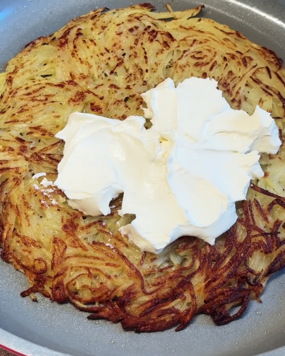 White cream cheese being dolloped and smoothed out across the hot crispy potato pancake.