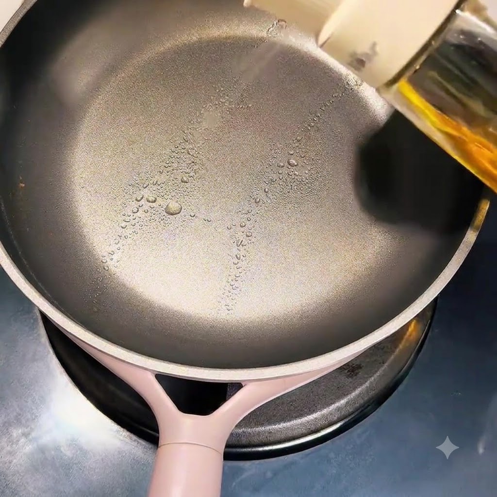 A hand spraying cooking oil onto the surface of a clean non-stick frying pan.