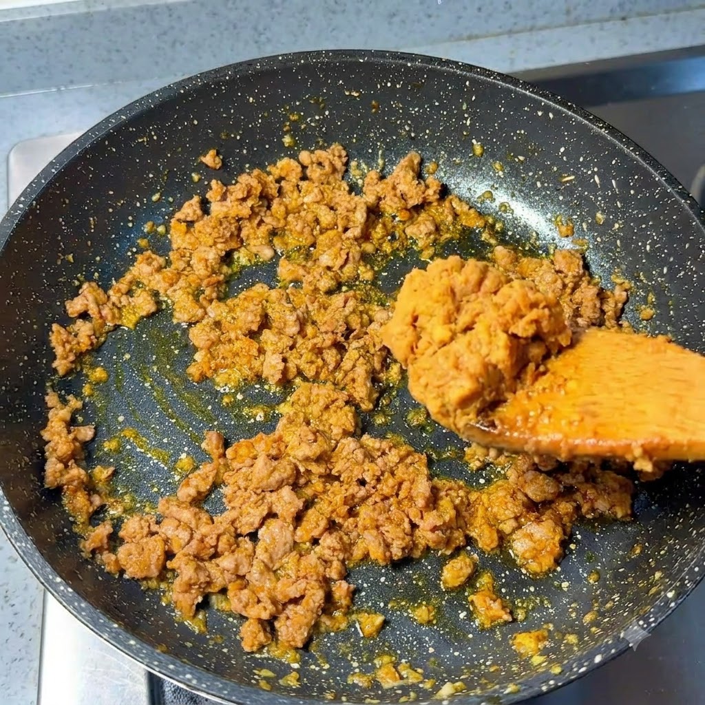 A spoonful of miso paste being added to the stir-fried minced pork in the pan.