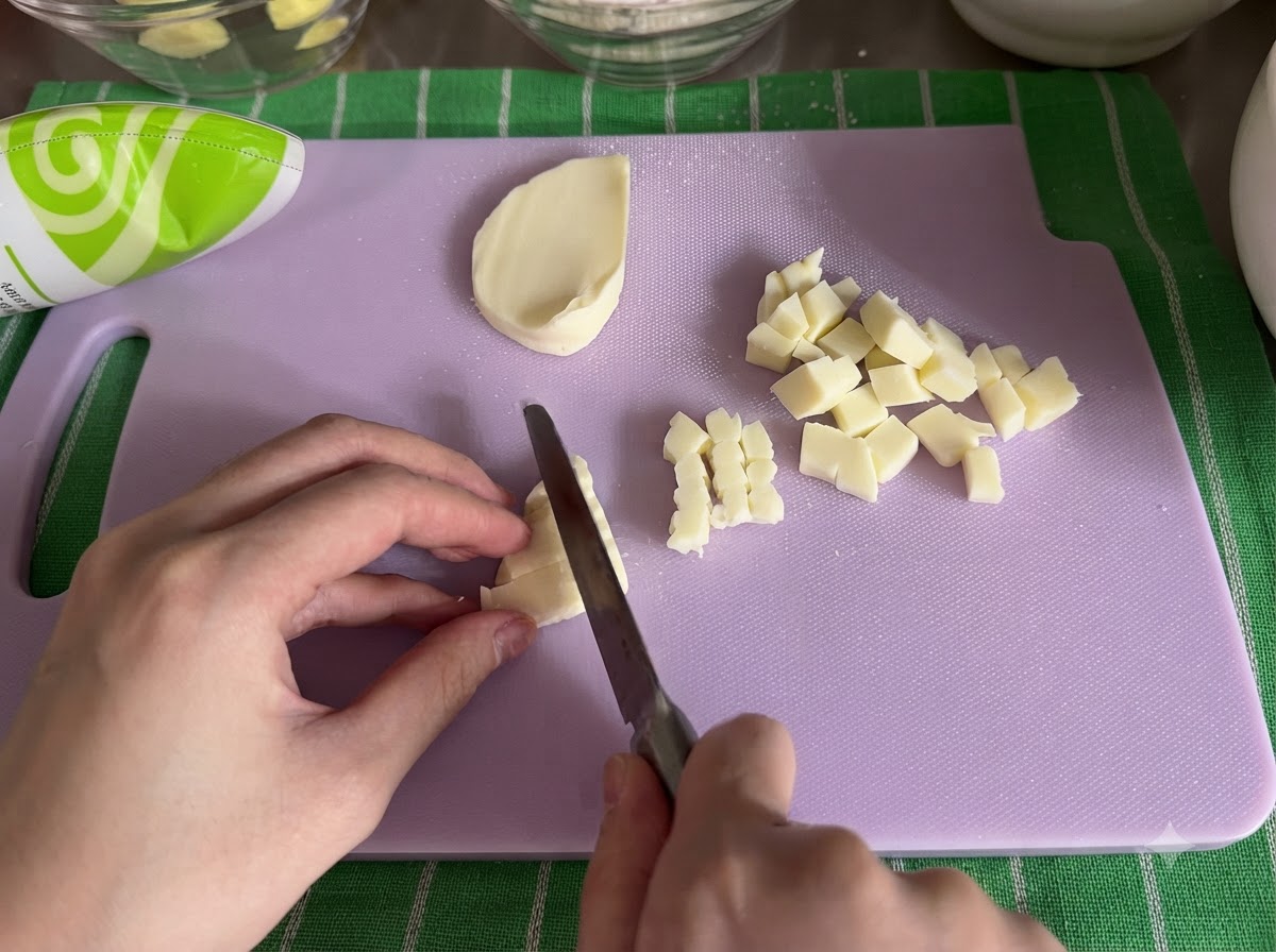 A block of mozzarella cheese being sliced into small cubes with a knife on a cutting board.