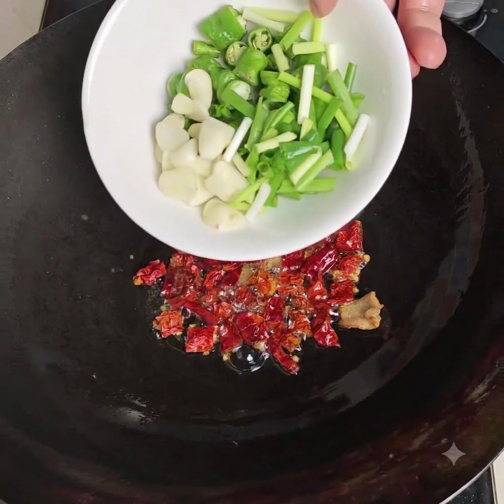 A white bowl pouring sliced green chilies, peeled garlic, and scallions over the toasted dried chilies in the wok.