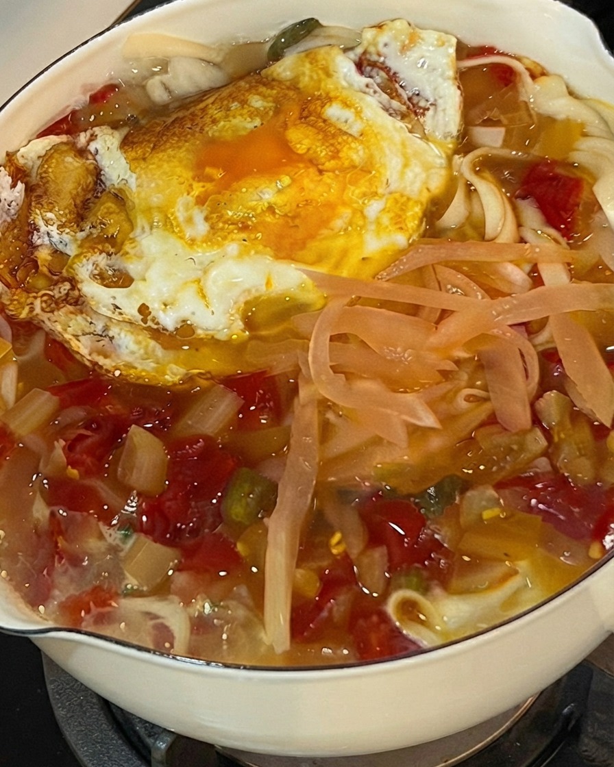 Fresh chopped cilantro being sprinkled on top of a bubbling pot of noodles, pickled cabbage, and a fried egg.