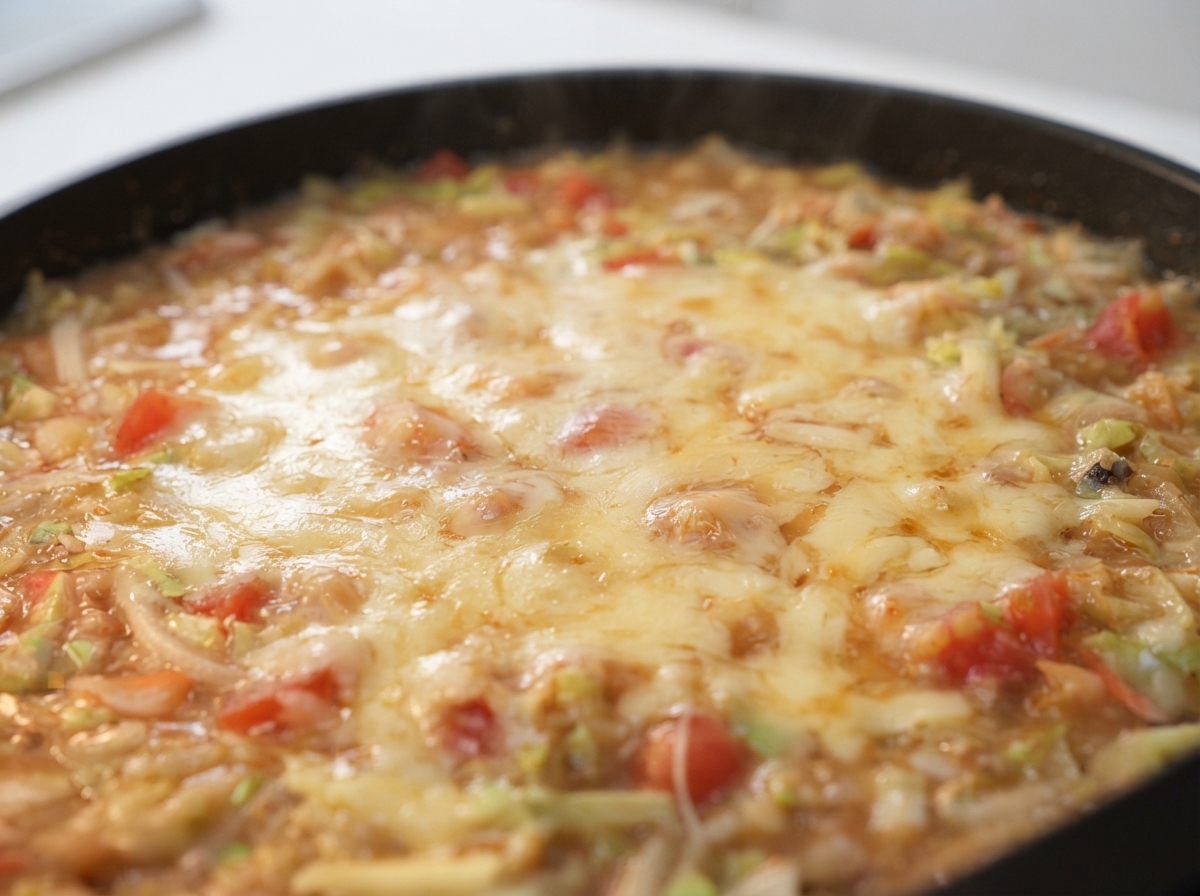 Shredded cheese melting over a bubbling mixture of batter and vegetables on a hot pan.