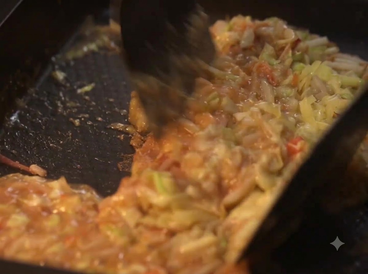 Spatulas mixing cooking vegetables and liquid batter on a hot plate.