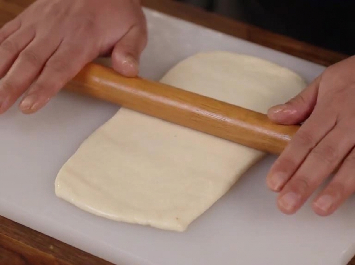 Hands using a wooden rolling pin to flatten a smooth piece of dough into a rectangle on a white cutting board.