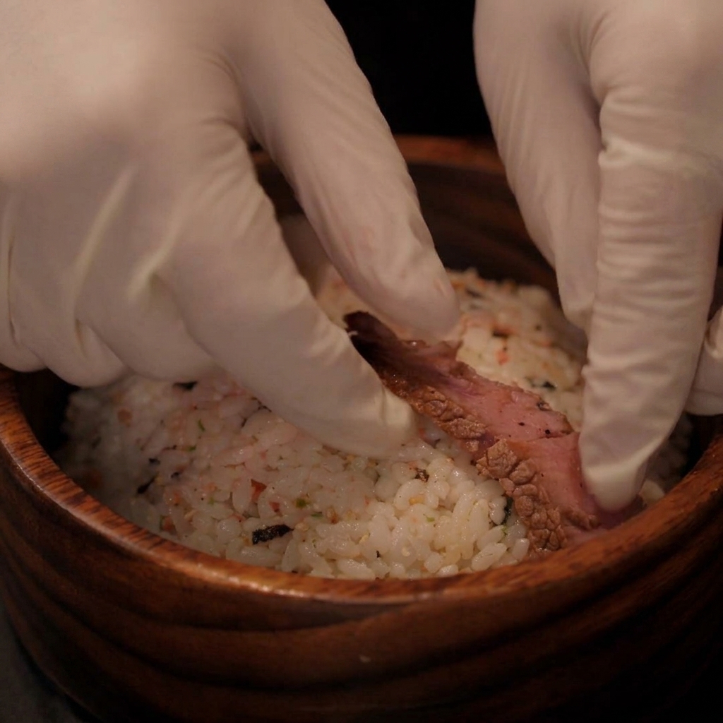 Gloved hands placing cooked steak slices onto a bowl of seasoned rice.