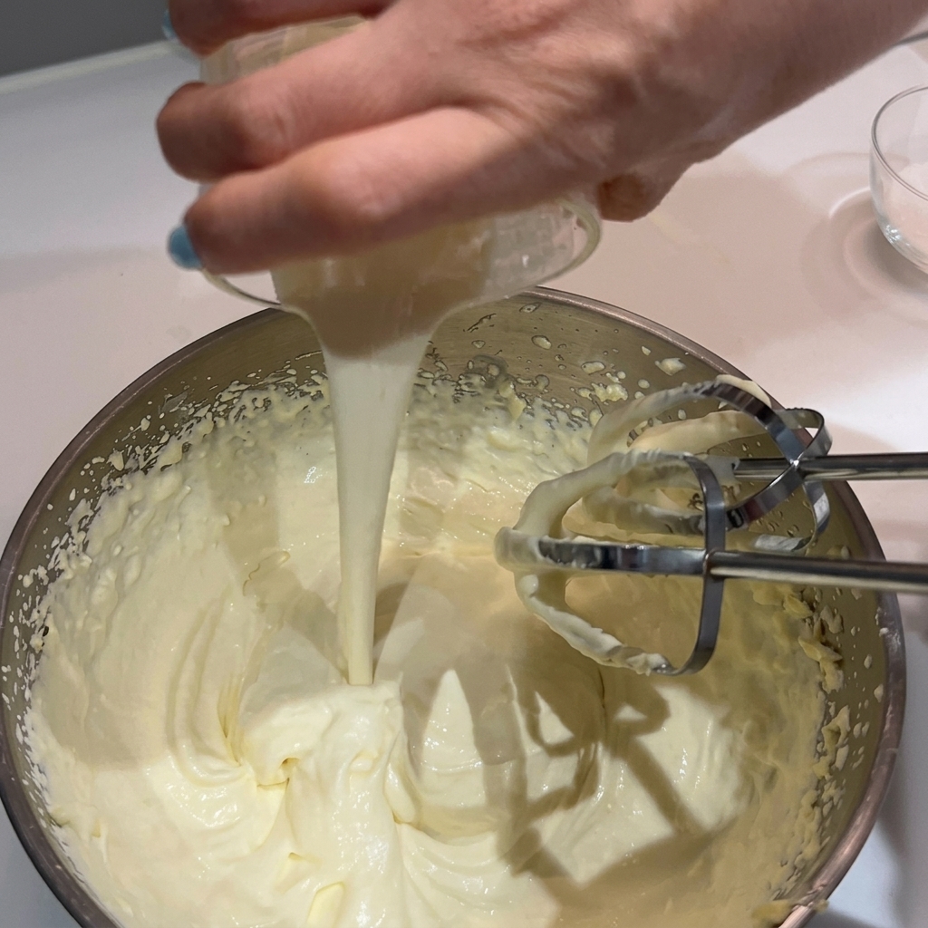 A hand pouring more cream into a bowl of whipped cream and butter with electric beaters.