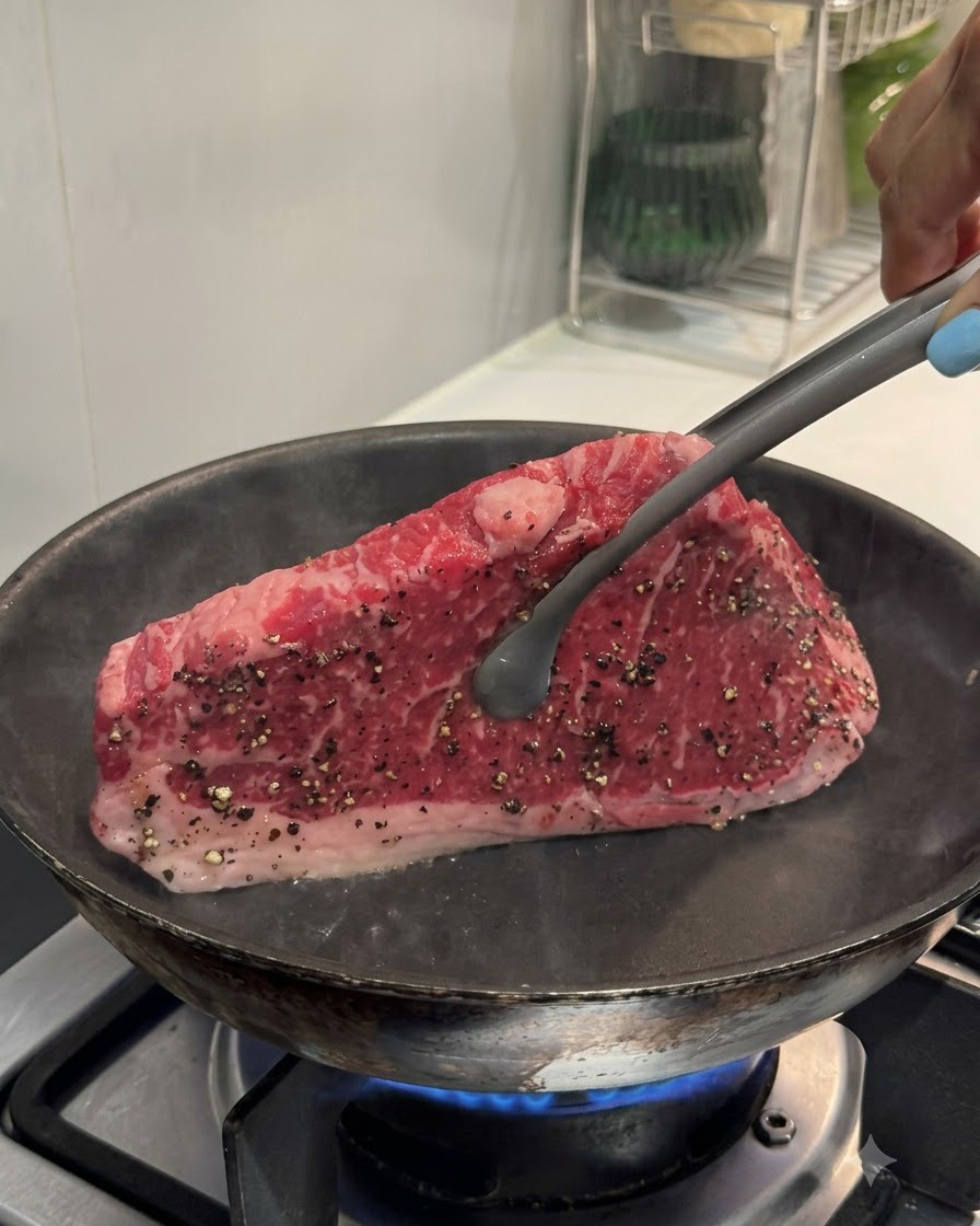 Tongs holding a steak upright against a hot pan to sear the edge.