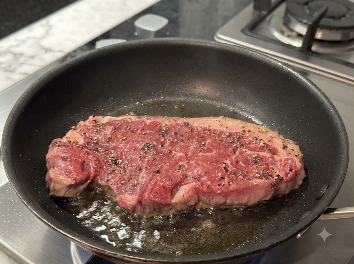 A steak frying in a hot pan, showing a browned crust forming on the surface.