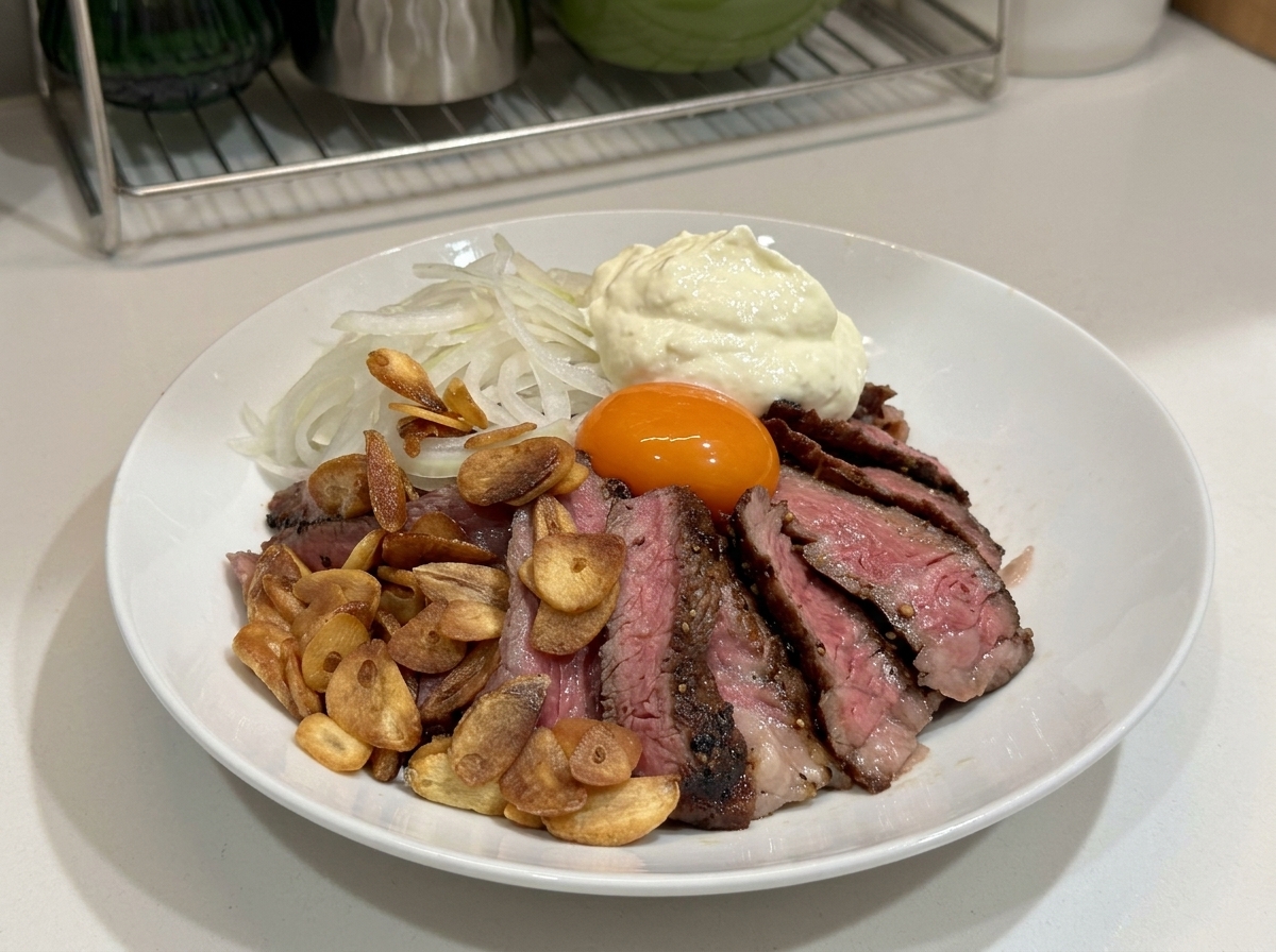 A white bowl filled with sliced steak, garlic chips, onions, an egg yolk, and a large dollop of whipped cream on top.