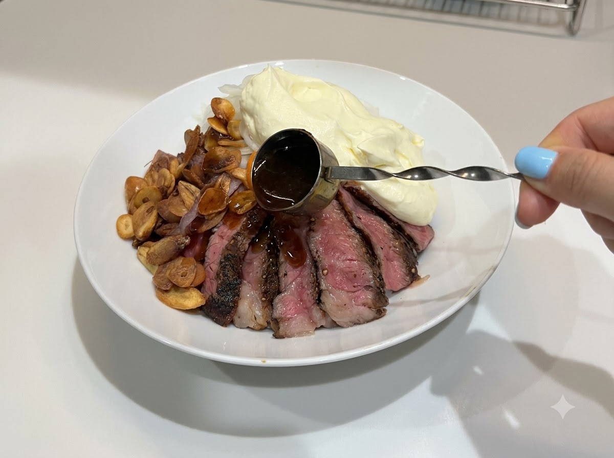 A hand using a small handled measure to drizzle dark, glossy teriyaki sauce over a completed steak rice bowl.
