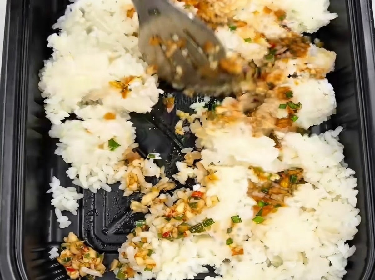 Rice being stirred and mixed with tongs inside a black takeout container alongside garlic sauce.