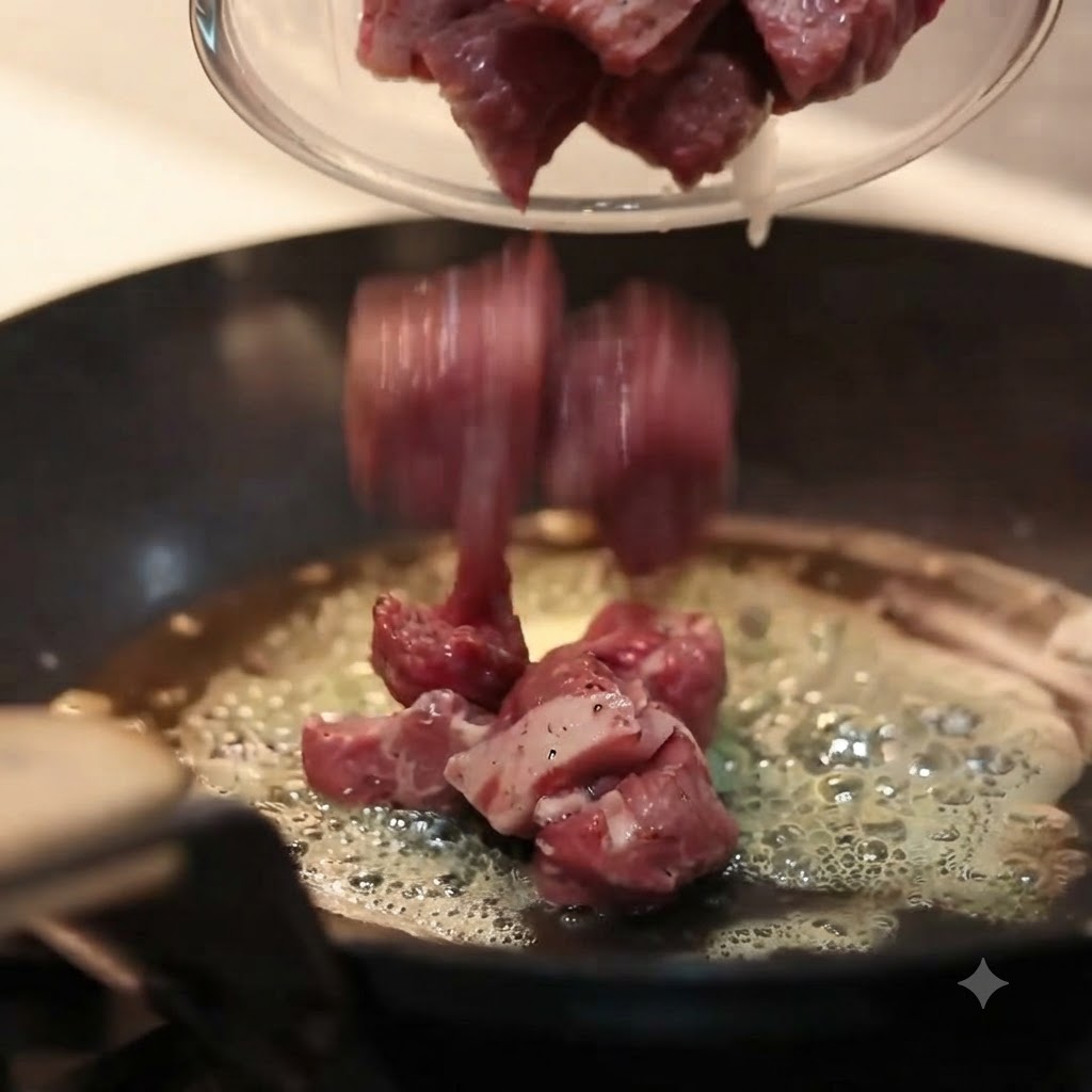 Pieces of seasoned raw beef being poured from a glass bowl into a hot, flat-bottomed pan filled with bubbling melted butter.