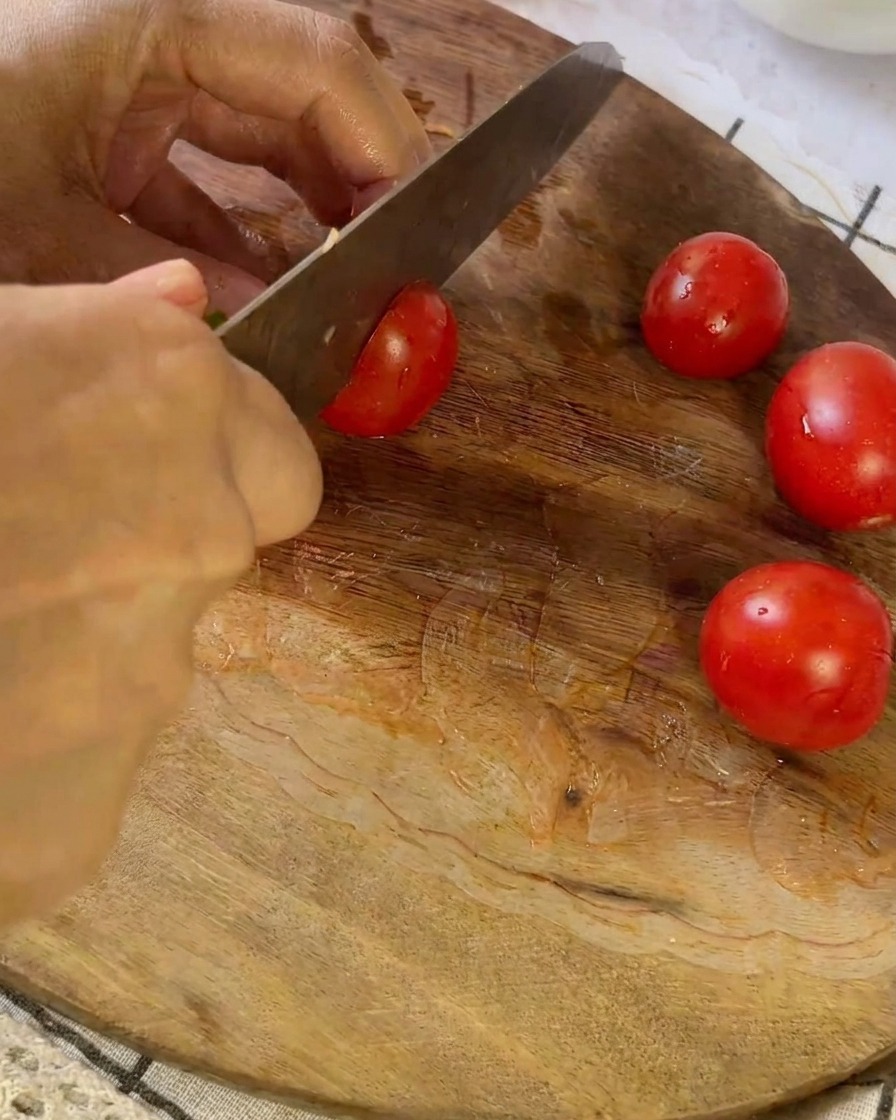 A knife slicing through a small red tomato on a wooden cutting board next to other halved tomatoes.
