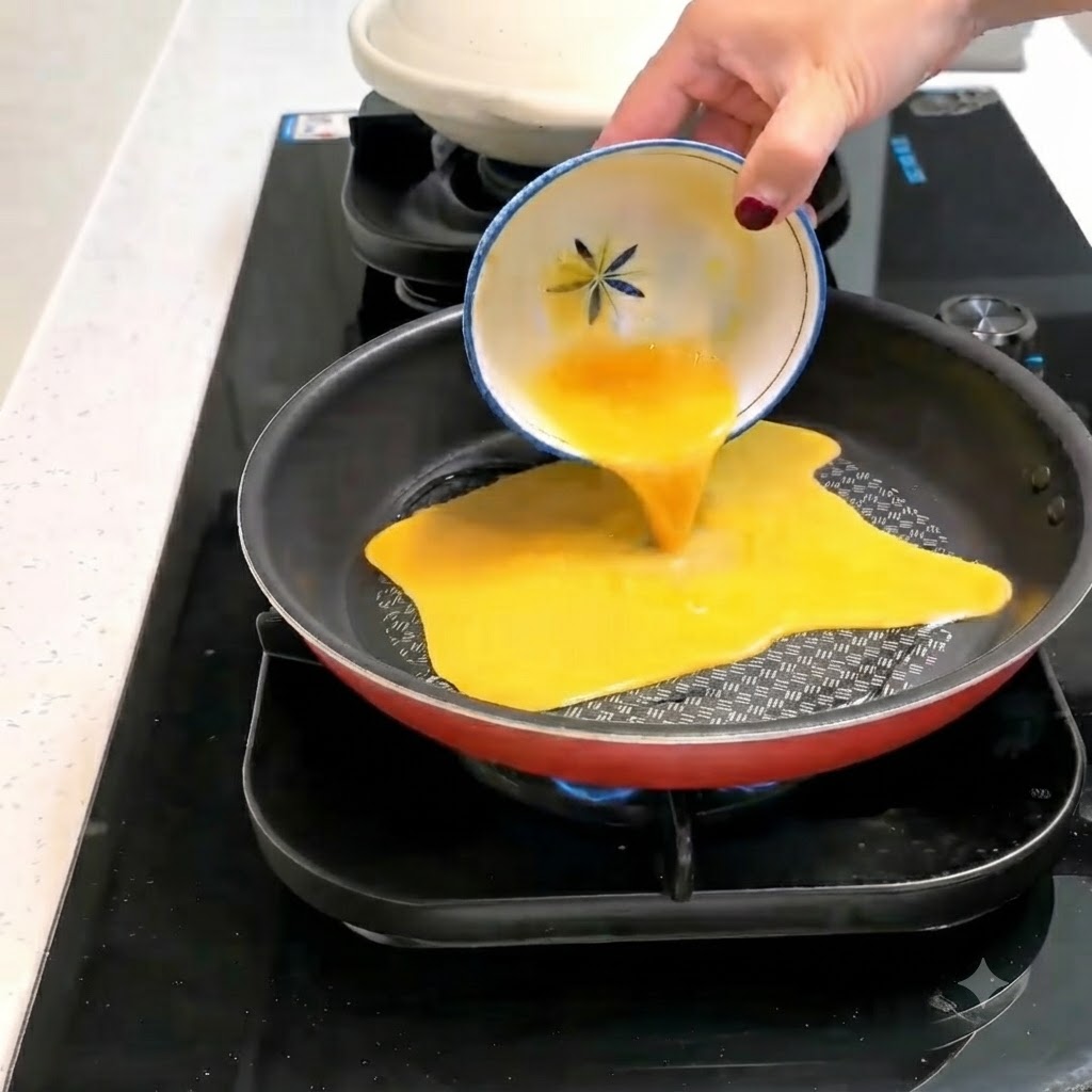 A beaten egg mixture being poured from a small patterned bowl into a hot, oiled frying pan.