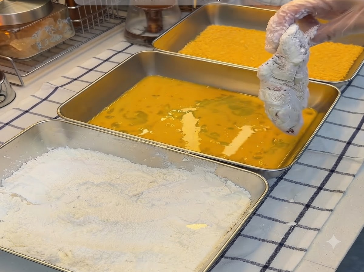 A starch-coated pork cutlet being dipped into a tray of beaten yellow eggs.