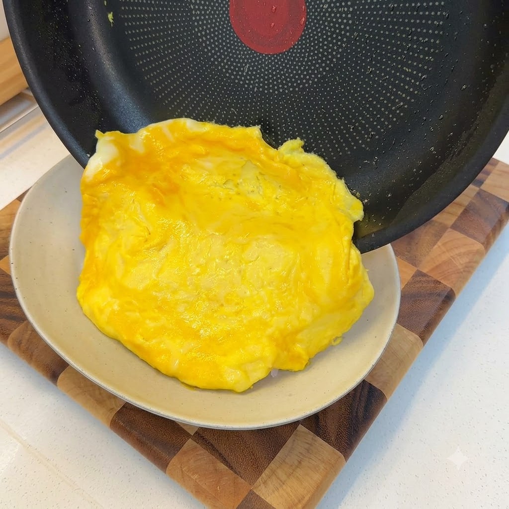 A soft cooked omelet sliding out of a frying pan and draping over a portion of white rice.