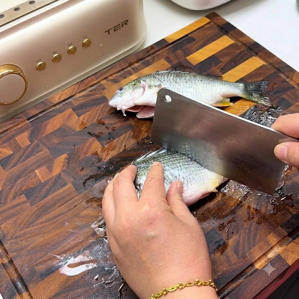 A hand using a large metal cleaver to make diagonal scores on the side of a fresh fish on a wooden cutting board.