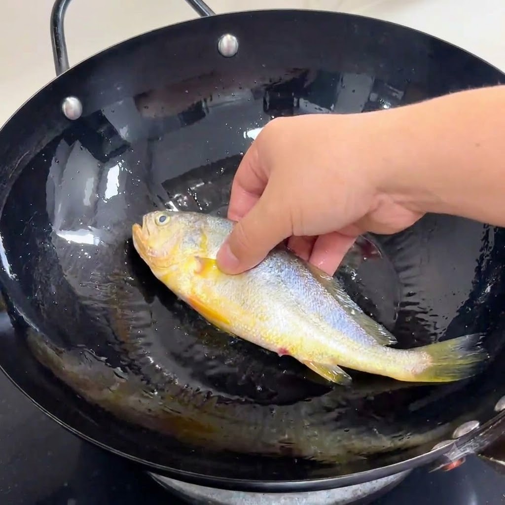 A hand gently placing a whole seasoned fish into a black wok filled with heated cooking oil.