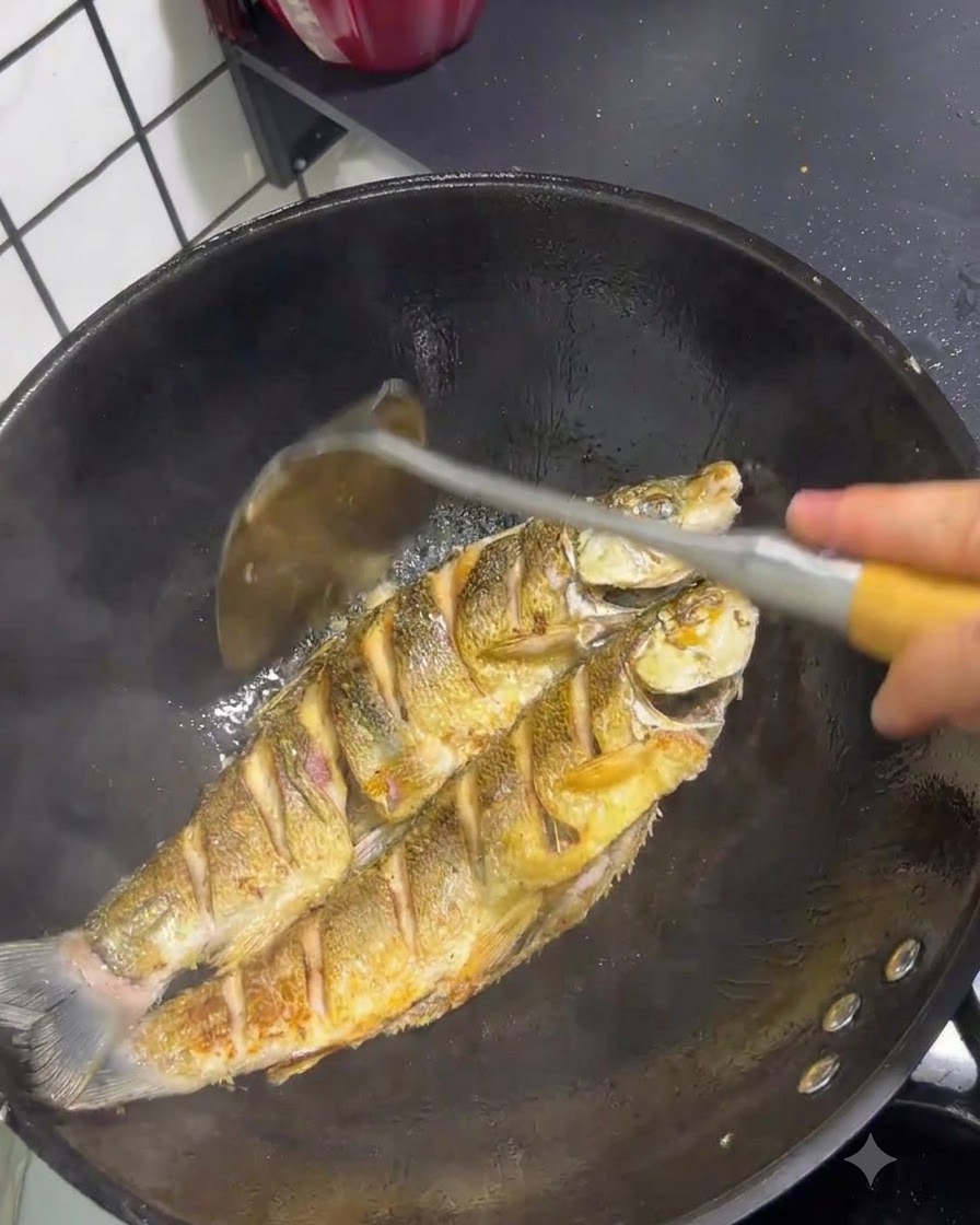 Two yellow tail fish frying in a wok with golden brown skin being managed by a spatula.