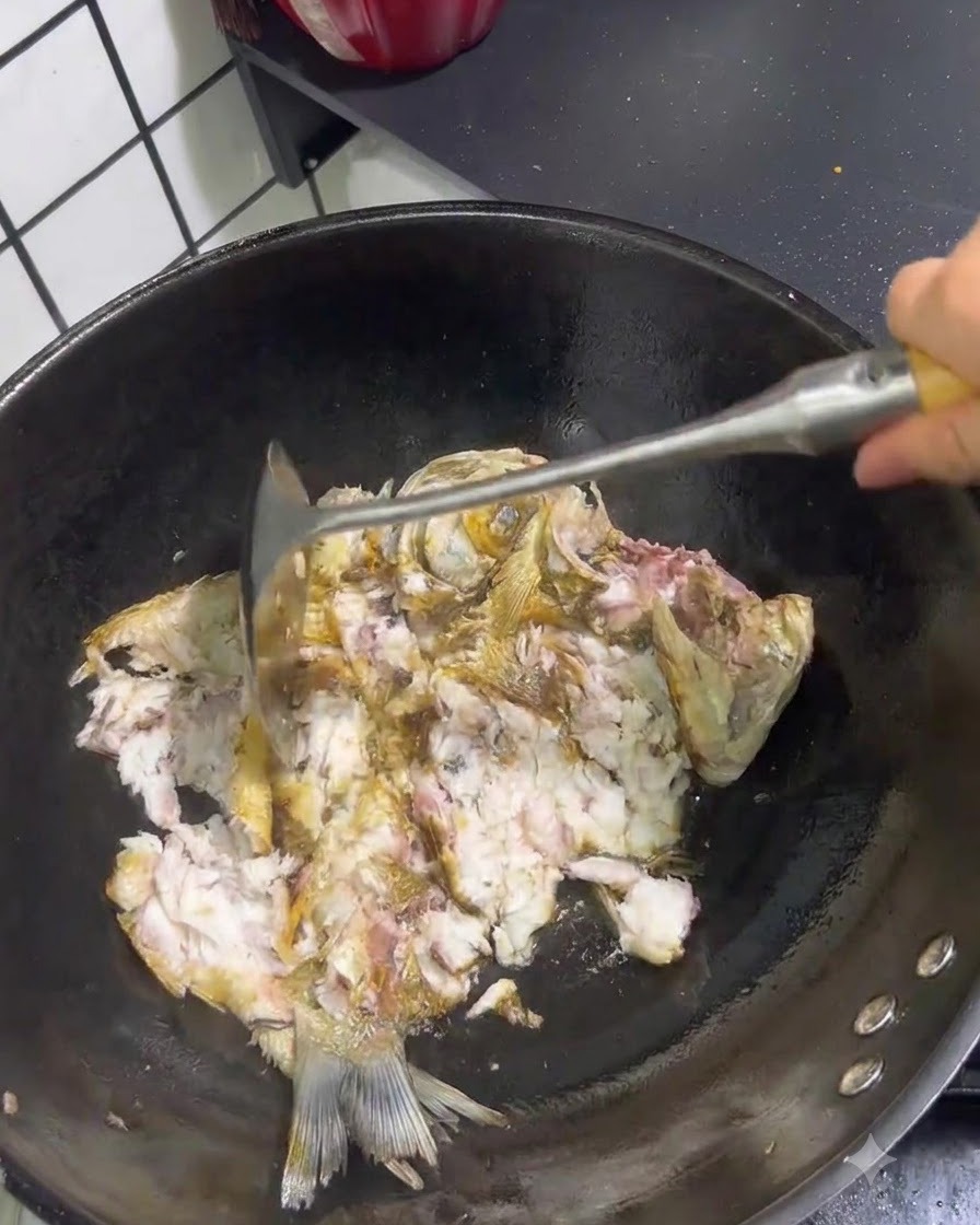 Fried fish being broken apart and crushed with a metal spatula inside a wok.
