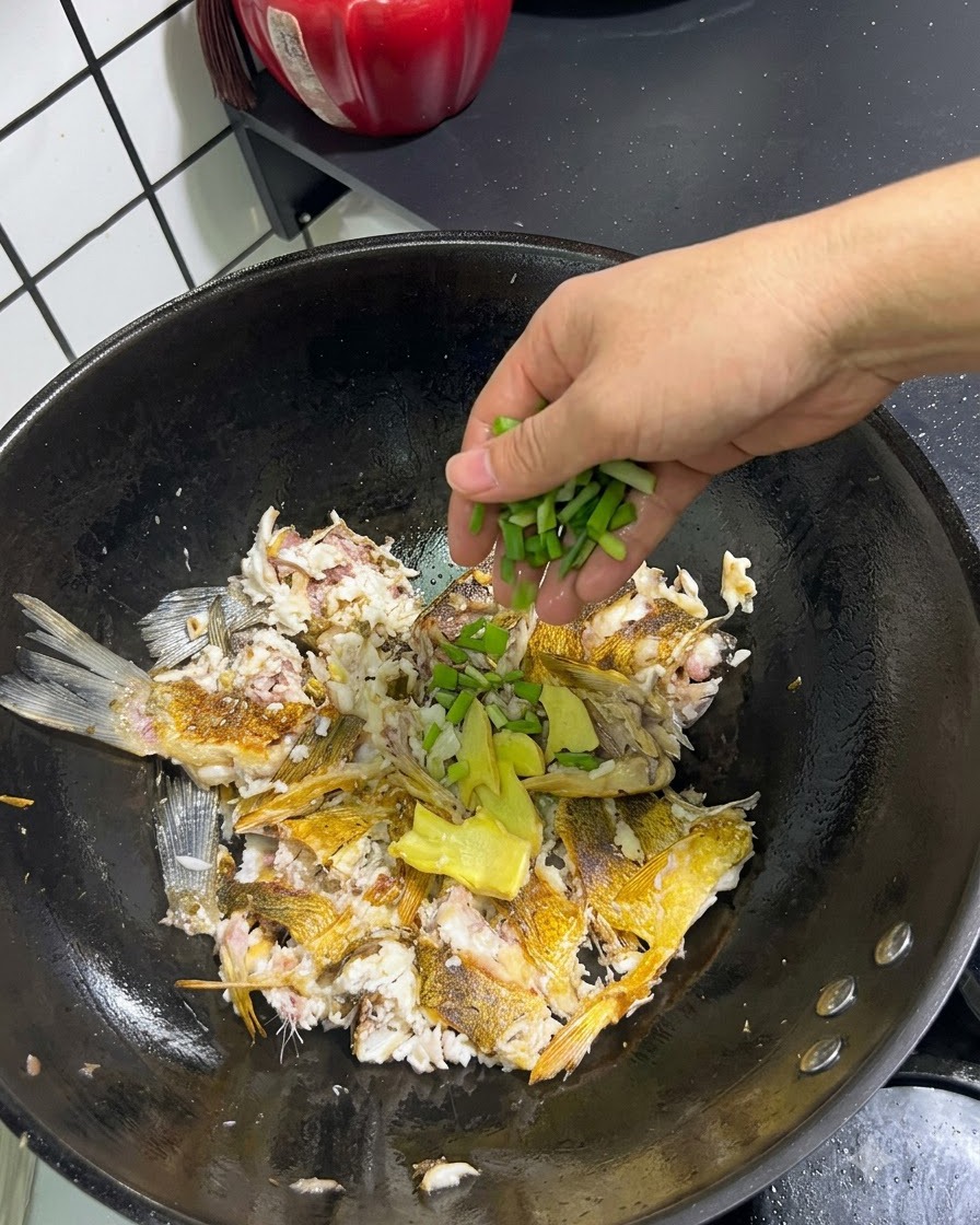 Fresh green onions and sliced ginger being added by hand over crushed fish in a wok.