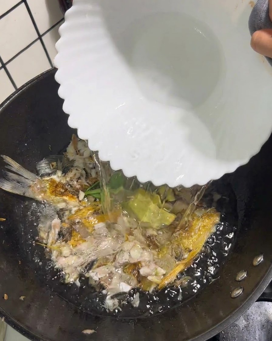 Clear boiling water being poured from a white bowl into a wok containing crushed fish and aromatics.