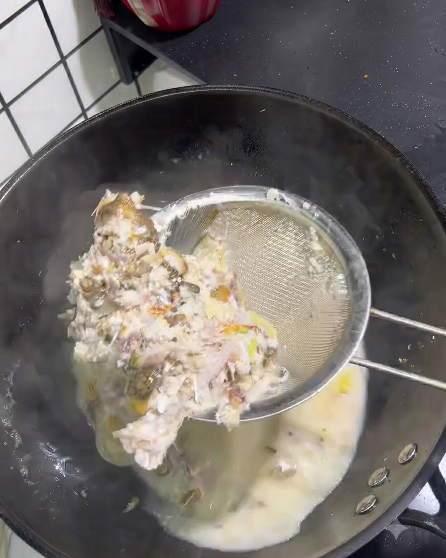 Solid fish bones and meat being scooped out of a milky broth with a wire mesh strainer.
