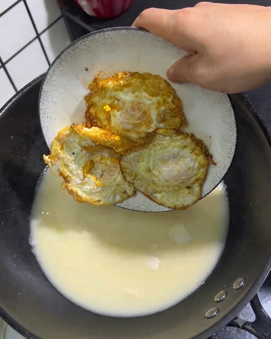 Three golden fried eggs being placed from a white plate into a pot of simmering soup.