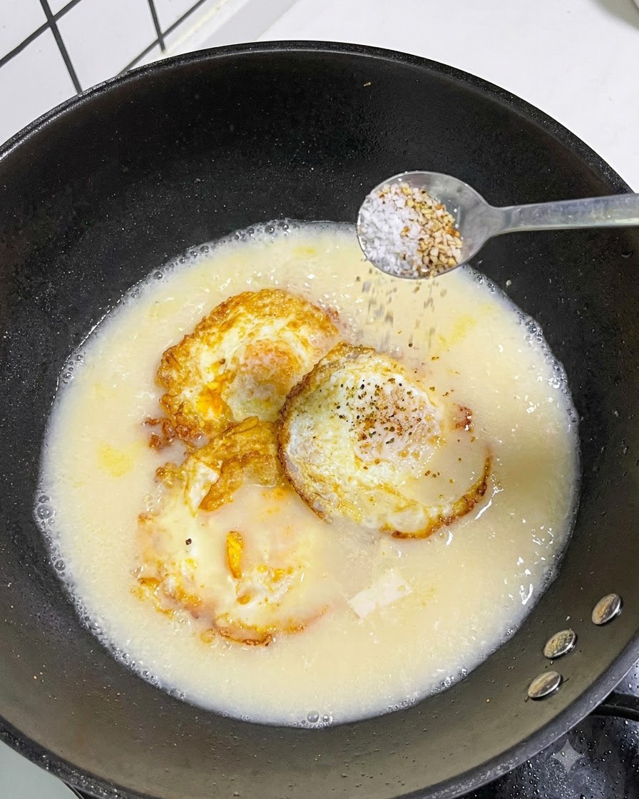 A spoonful of salt and white pepper being sprinkled from a spoon into a pot of soup.