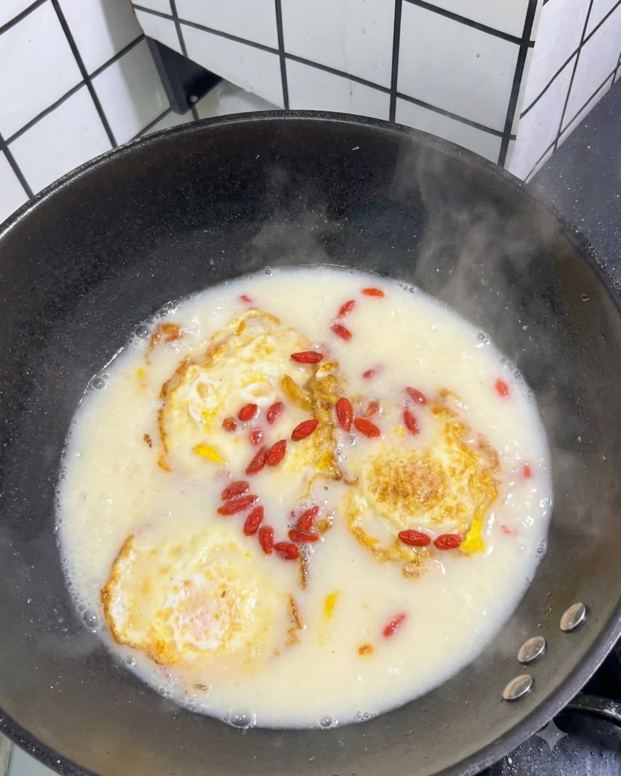 Dried red goji berries scattered across the surface of a simmering white soup with fried eggs.