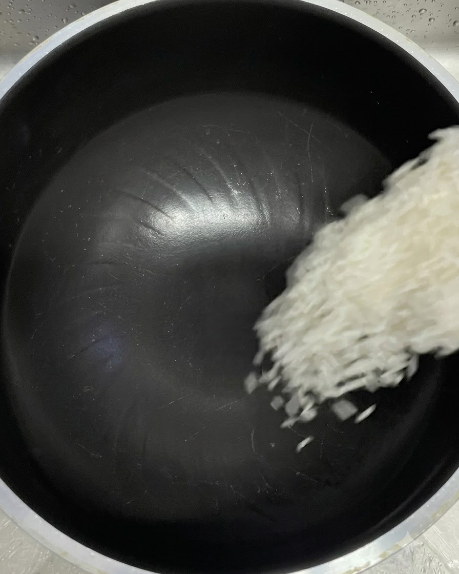 White rice grains falling from a measuring cup into a black rice cooker inner pot.