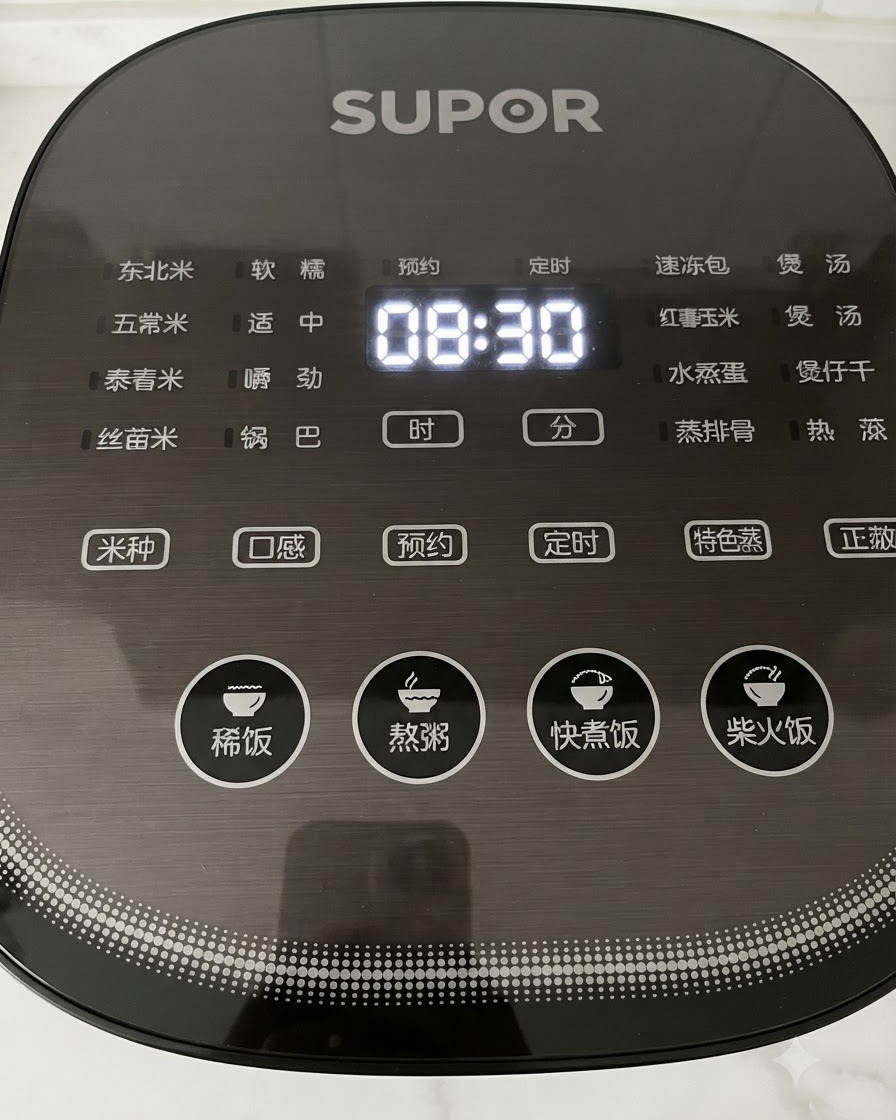 A close-up view of a hand pressing buttons on a black digital rice cooker control panel displaying time.
