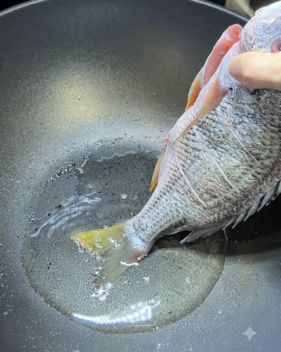 A hand holding a whole raw yellow snapper by the upper body as its tail touches the hot oil in a wok.