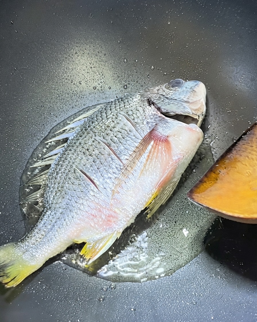 A whole yellow snapper frying in a wok with a wooden spatula positioned nearby for flipping.