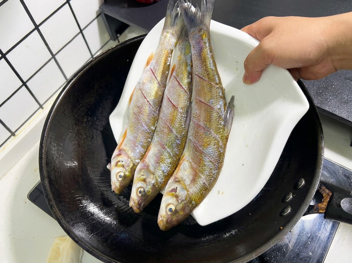 Three whole, scored raw fish being slid off a white plate into a black wok with hot oil.