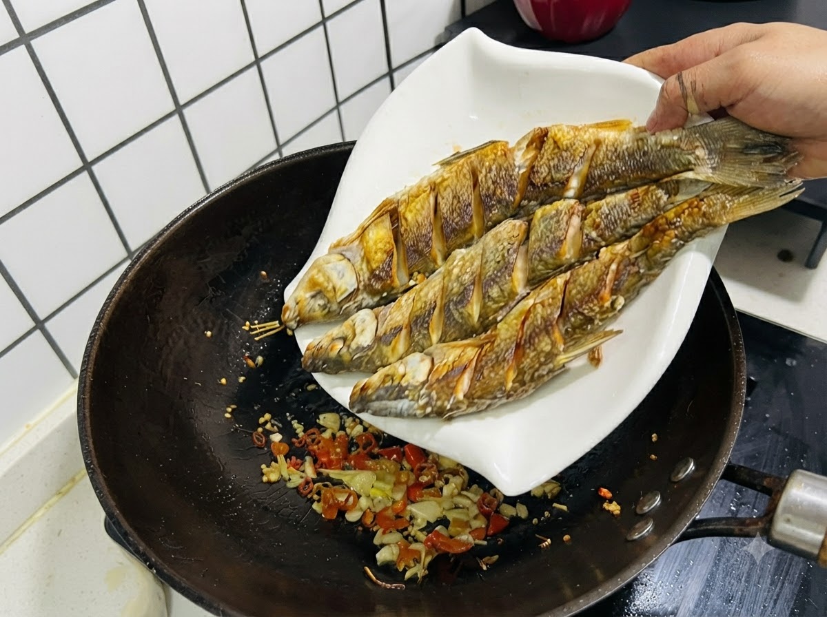 Three fried fish being slid from a white plate back into a wok containing sautéed garlic, ginger, and chilies.