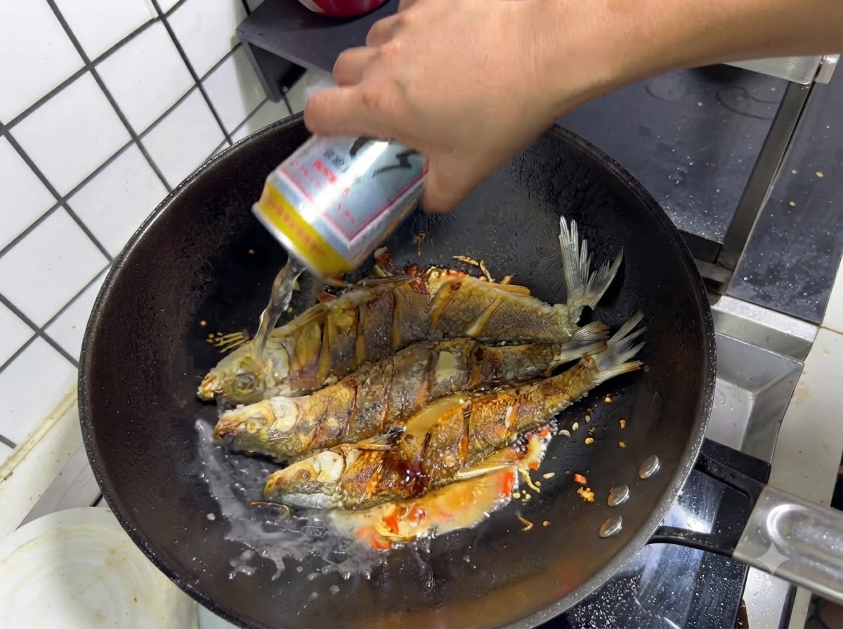 A hand pouring a can of beer into a wok over the seasoned fried fish and sautéed aromatics.