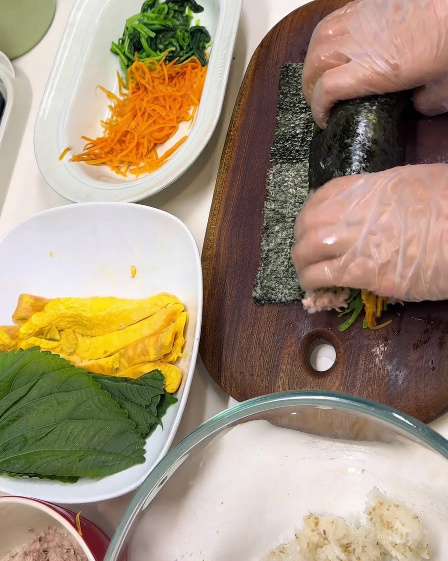 Hands wearing clear gloves wrapping ingredients inside a sheet of nori on a wooden cutting board.