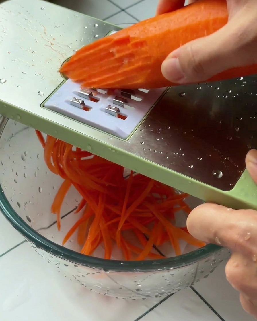 A hand holding a peeled carrot against a green-framed grater, shredding it into a glass bowl.