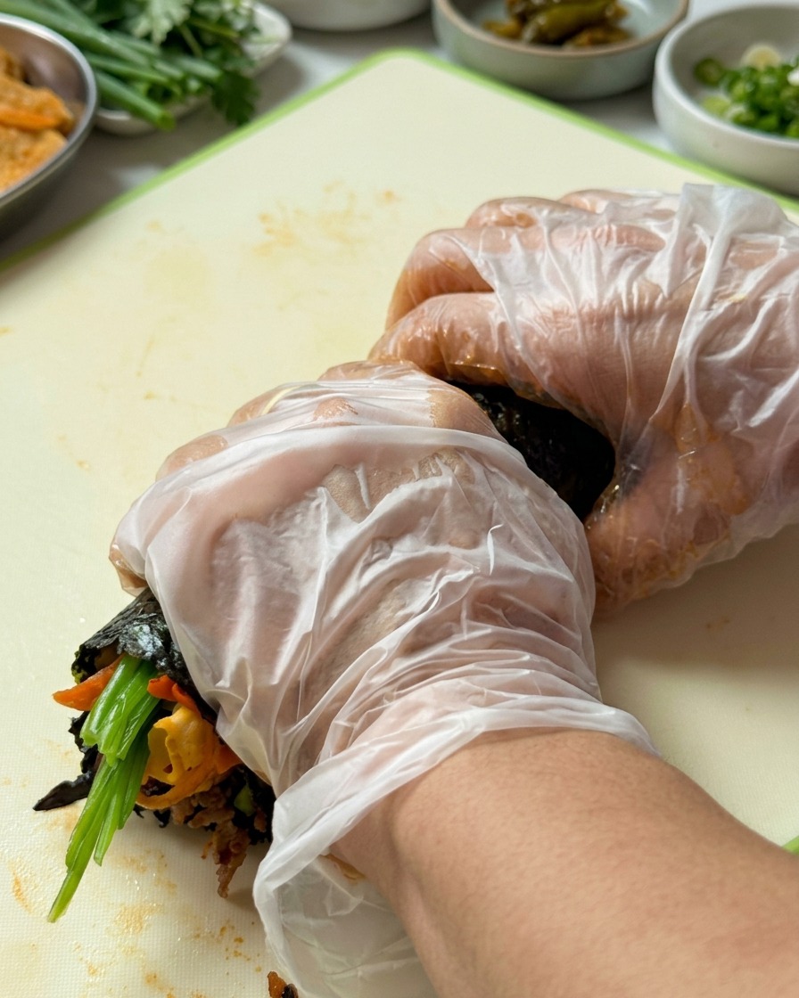 Gloved hands firmly rolling the gimbap from the bottom edge upwards.
