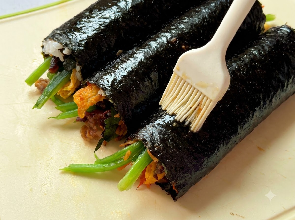A close-up view of sesame oil being brushed onto three rolled gimbap logs placed on a cutting board.