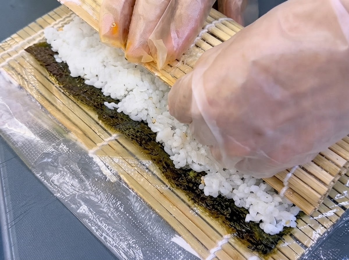 Gloved hands lifting a bamboo mat to tightly roll seaweed and fillings into a cylinder.