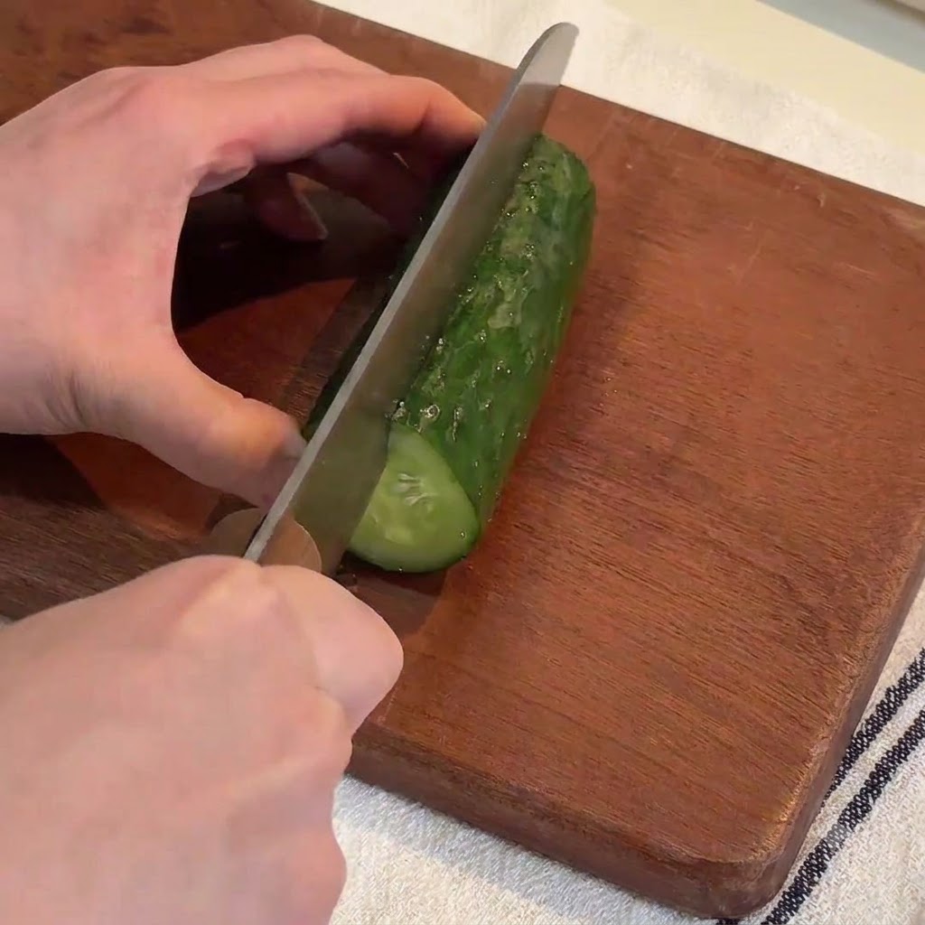 Hands using a large knife to slice a fresh green cucumber into long strips on a wooden cutting board.