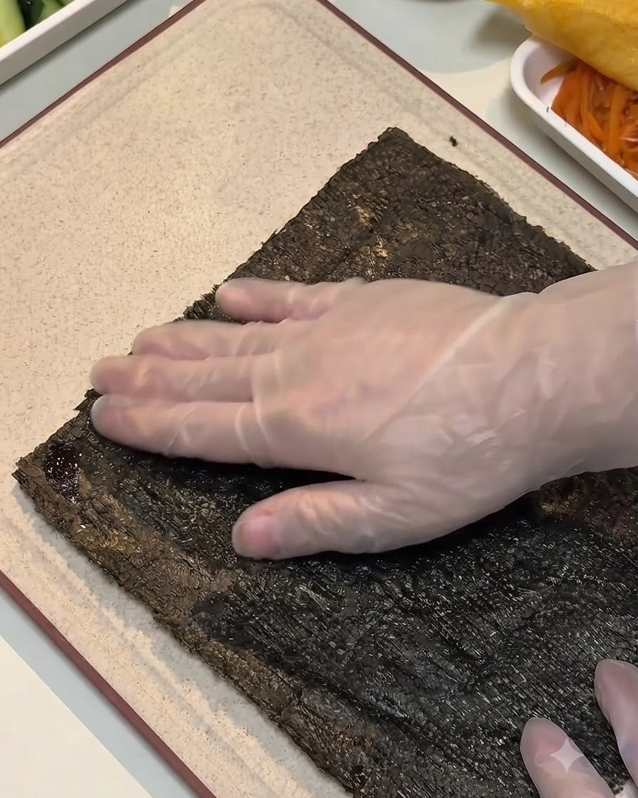 A gloved hand pressing down on the edge of a dampened seaweed sheet on a cutting board.