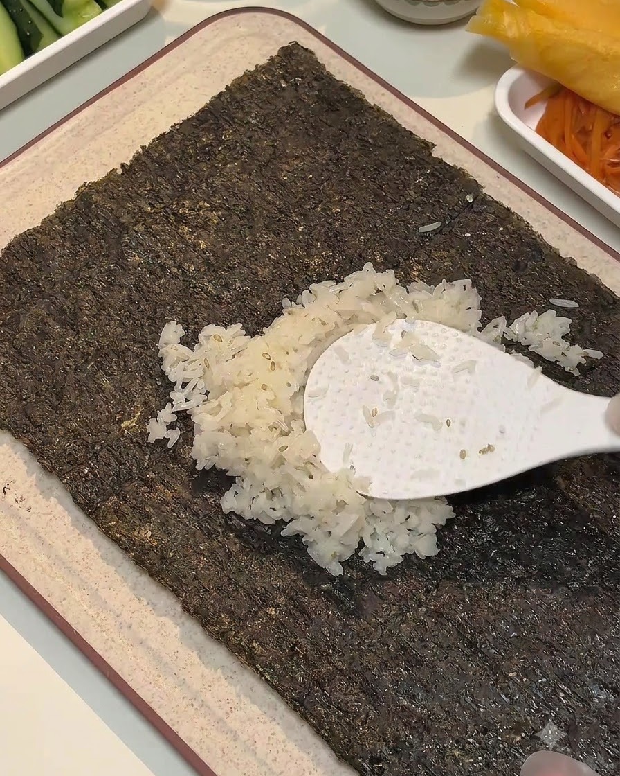 A white rice paddle spreading seasoned rice across the center of a seaweed sheet.