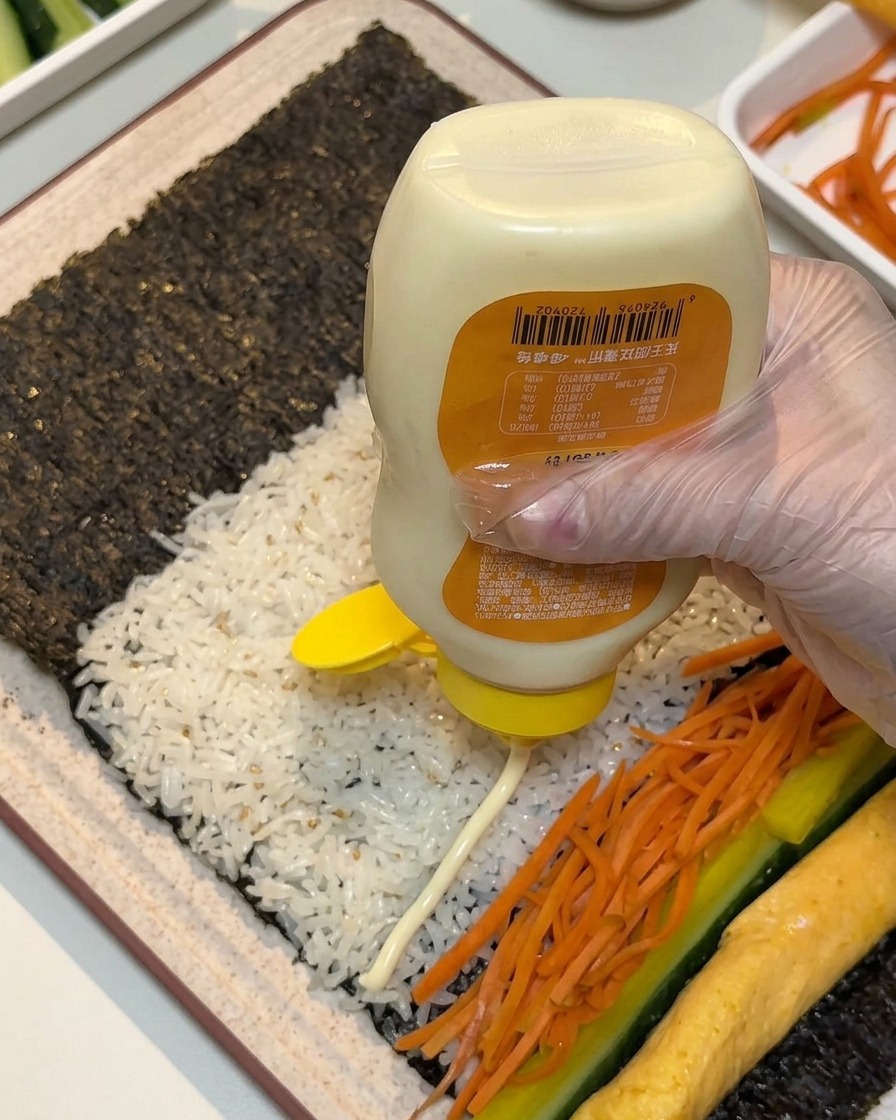 A gloved hand squeezing lines of white mayonnaise from a bottle across the laid-out fillings on seaweed and rice.