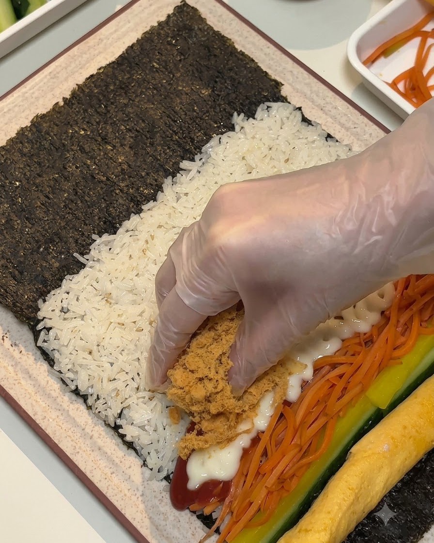 A gloved hand placing brown fluffy meat floss and luncheon meat strips over the fillings on the seaweed sheet.