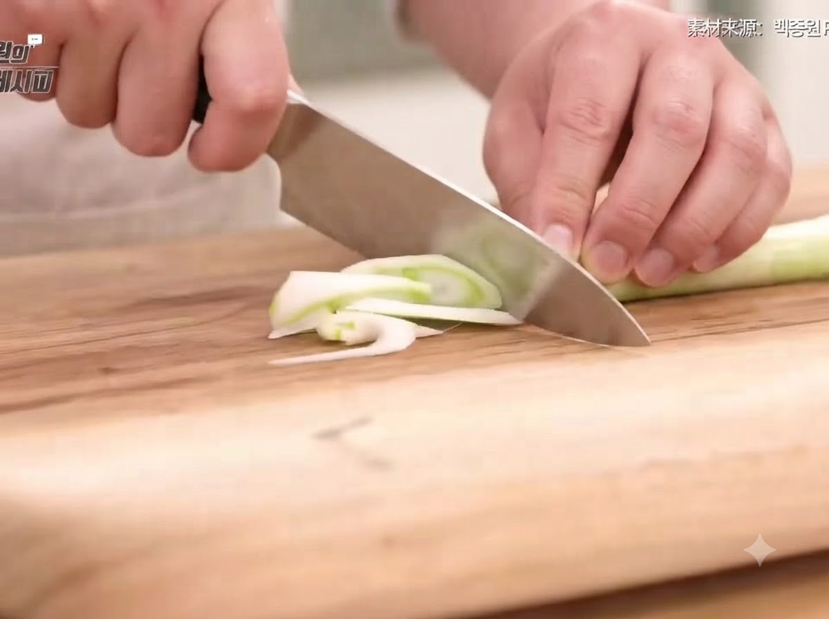 Green onion, onion, carrot, and chili peppers being cut into thin slices and shreds on a wooden cutting board with a knife.