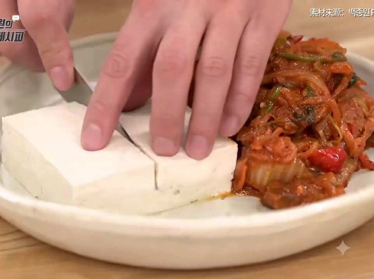 A hand using a knife to slice blanched tofu neatly on a plate next to a mound of savory stir-fried kimchi and pork.